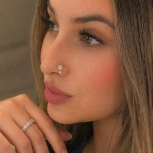 May include: A close-up of a woman's face, featuring a gold nose stud with a floral design and a dangling charm. The woman also wears a ring with multiple small, clear stones. Her skin has a warm, pinkish tone, and her hair is long and light brown.