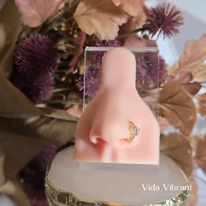 May include: A close-up of a nose piercing with a gold setting and clear stones. The piercing is on a model's nose.