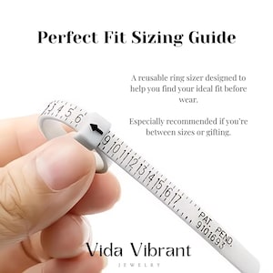 以下が含まれることがあります： 「Perfect Fit Sizing Guide」と「Vida Vibrant Jewelry」の文字が入った、黒い目盛りの白いリングサイザー。購入前にリングのサイズを測るためのものです。手で持っています。