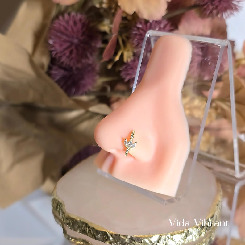 May include: A gold nose ring with a small, clear crystal design. The ring is shown on a model's nose.