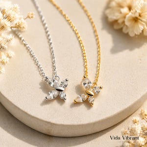 Puede incluir: Dos collares con colgante de mariposa, uno plateado y otro dorado, cada uno con detalles de piedras preciosas transparentes. Los collares se exhiben sobre una superficie neutra con flores secas, mostrando el diseño delicado y el brillo de la joyería.