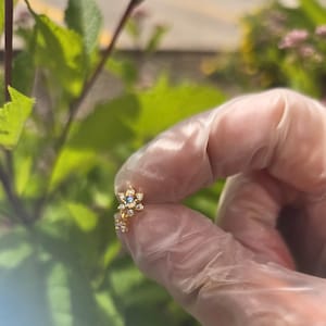 May include: A delicate gold-coloured floral earring featuring a central blue gemstone and clear crystal accents. The earring is held in a gloved hand, set against a background of green leaves and pink flowers.