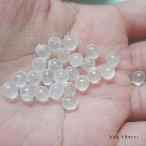 Puede incluir: Primer plano de una mano que sostiene varios topes de pendientes redondos y translúcidos. Los topes son pequeños, con una base ligeramente aplanada y una parte superior redondeada. El color es blanco lechoso y el material parece ser plástico flexible.