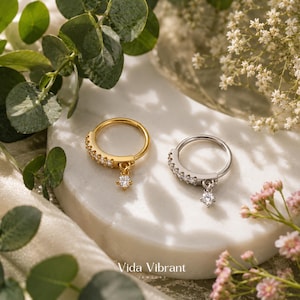 May include: Two circular hoop rings, one gold-toned and one silver-toned, each featuring a row of small clear stones along the outer edge and a single dangling clear stone charm. The rings are displayed on a smooth, white, rounded surface.