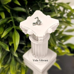 May include: A silver nose ring with a floral design and clear gemstones is displayed on a white decorative pedestal. The pedestal has a classical column design and the words "Vida Vibrant Jewelry" are printed on the base.