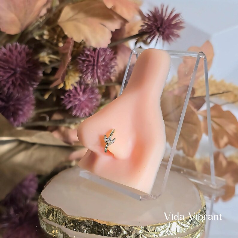 May include: A close-up of a nose piercing with a gold lightning bolt design featuring small clear stones. The piercing is on a model's nose and is displayed on a clear acrylic stand.
