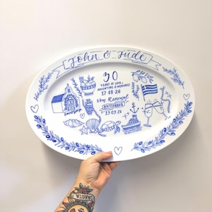 May include: An oval white ceramic platter featuring blue ink illustrations of a church, a sea turtle, a boat, a Greek flag, and champagne flutes. The text reads John & Jude, 30 Years of Love, Adventure & Memories, 17.08.26, Von Renewal, Antiparos, 19.09.26.
