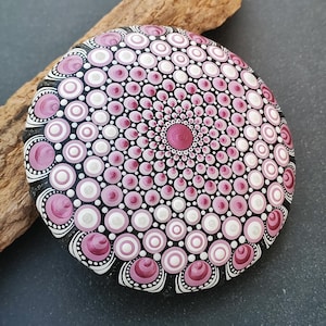 May include: A round stone painted with a mandala pattern featuring concentric circles in shades of magenta, light pink, and white against a black background. The design radiates from a central magenta dot, with intricate dot work throughout.