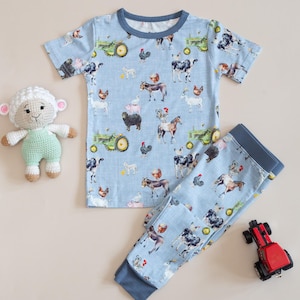 Op de afbeelding: Een lichtblauwe pyjamaset met een boerderijdierenprint, waaronder koeien, kippen en tractoren. De set bestaat uit een top met korte mouwen en een broek. Een gehaakt schaap en een rode speelgoedtrekker zijn ook aanwezig.