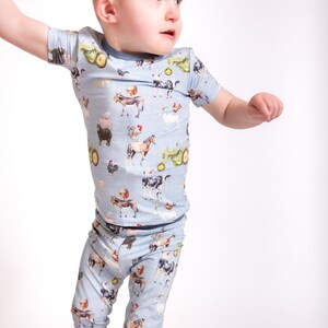 Op de afbeelding: Lichtblauw kinderpyjamaset met een boerderijdierenprint. De set bevat een top met korte mouwen en een bijpassende broek. De print toont koeien, kippen, paarden, tractoren en andere boerderij-elementen. De pyjama's hebben donkerblauwe biezen.