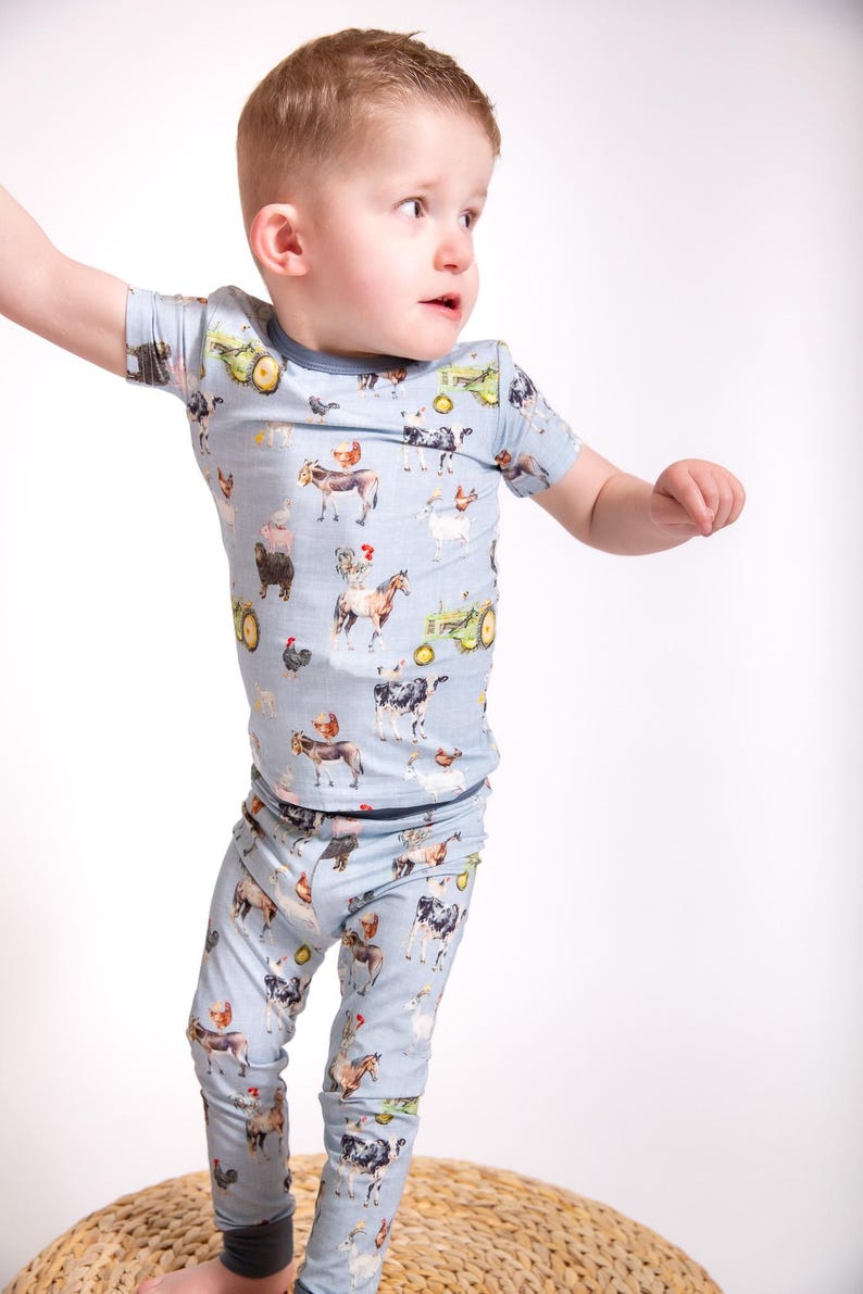 Op de afbeelding: Lichtblauw kinderpyjamaset met een boerderijdierenprint. De set bevat een top met korte mouwen en een bijpassende broek. De print toont koeien, kippen, paarden, tractoren en andere boerderij-elementen. De pyjama's hebben donkerblauwe biezen.