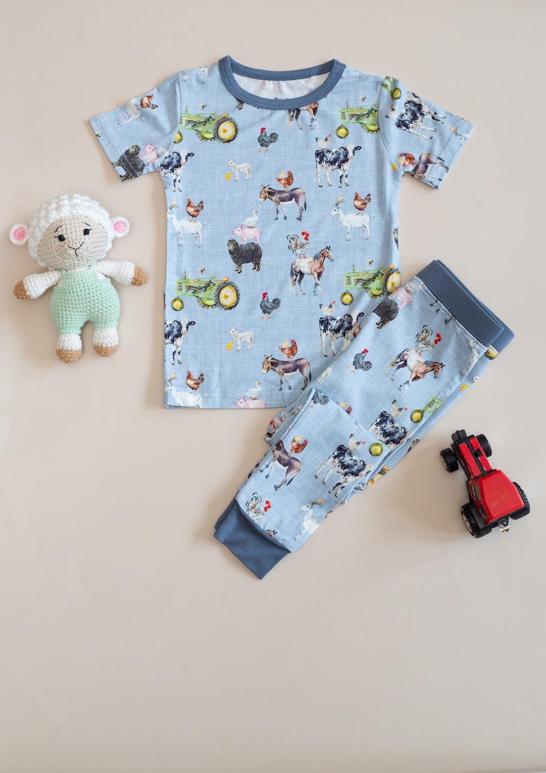Op de afbeelding: Een lichtblauwe pyjamaset met een boerderijdierenprint, waaronder koeien, kippen en tractoren. De set bestaat uit een top met korte mouwen en een broek. Een gehaakt schaap en een rode speelgoedtrekker zijn ook aanwezig.
