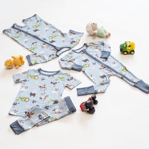 Op de afbeelding: Lichtblauwe baby pyjama's en een tweedelige set met een boerderijdierenprint. De pyjama's hebben een lange rits en blauwe biezen. Een gele speelgoedeend, een groen en wit schaap, een groene tractor en een rode speelgoedtruck staan ook op de foto.