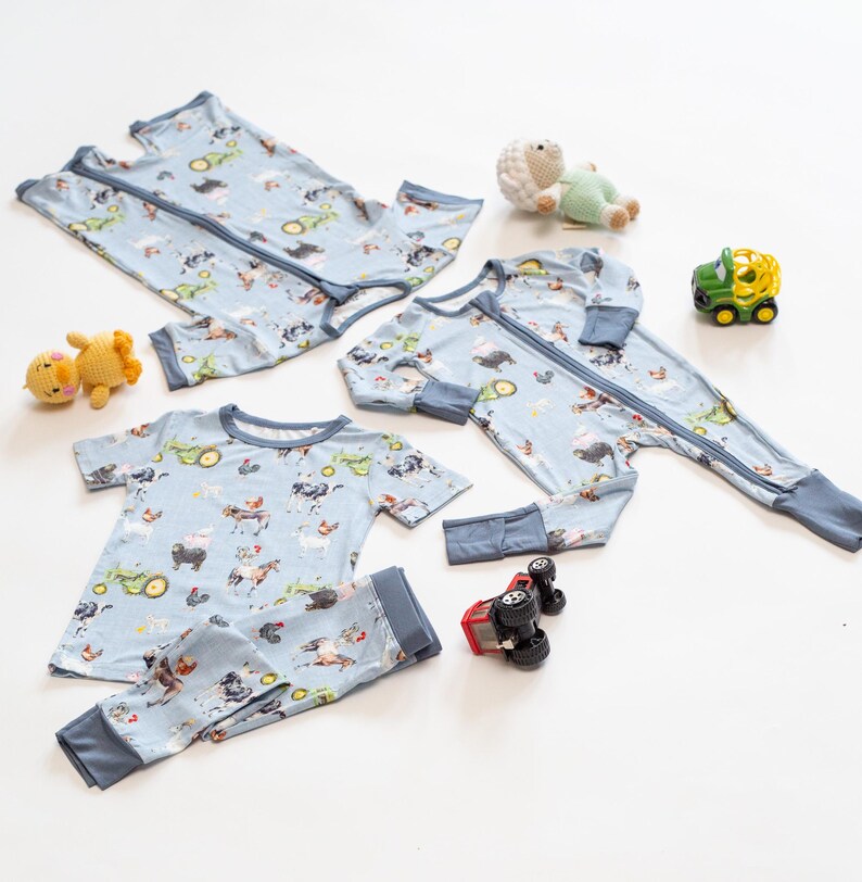 Op de afbeelding: Lichtblauwe baby pyjama's en een tweedelige set met een boerderijdierenprint. De pyjama's hebben een lange rits en blauwe biezen. Een gele speelgoedeend, een groen en wit schaap, een groene tractor en een rode speelgoedtruck staan ook op de foto.