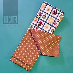 May include: Two fabric napkins, one with a white background and a pattern of lemons, berries, flowers, and plaid squares, and the other with a brown background and white polka dots.