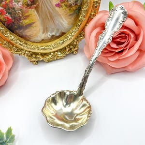 May include: A silver serving spoon with an ornate handle and a scalloped bowl. The spoon is resting on a white surface with a pink rose and a gold picture frame in the background.