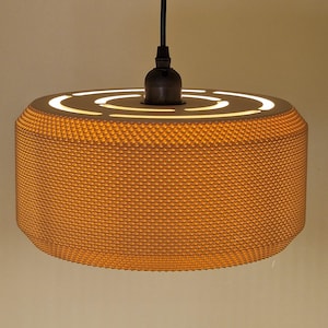 May include: A contemporary pendant light with a cylindrical lampshade. The shade is a textured, warm orange, with concentric illuminated circles on top. It hangs from a black cord and fixture, offering a modern design. The light would suit a room.
