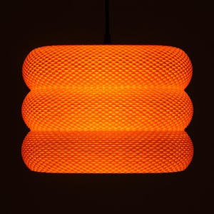 May include: An orange, textured pendant light. The light has a cylindrical shape with three stacked sections. The surface has a repeating diamond pattern. The light is suspended from a black cord against a dark background.