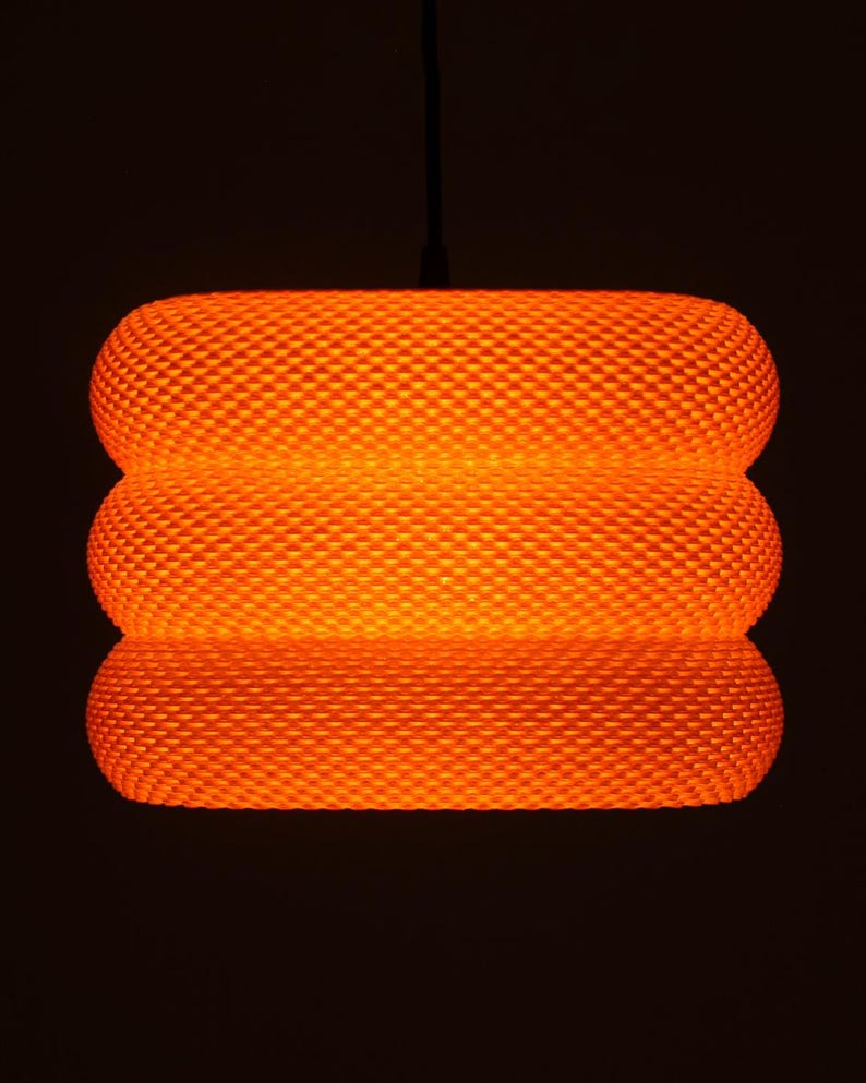 May include: An orange, textured pendant light. The light has a cylindrical shape with three stacked sections. The surface has a repeating diamond pattern. The light is suspended from a black cord against a dark background.