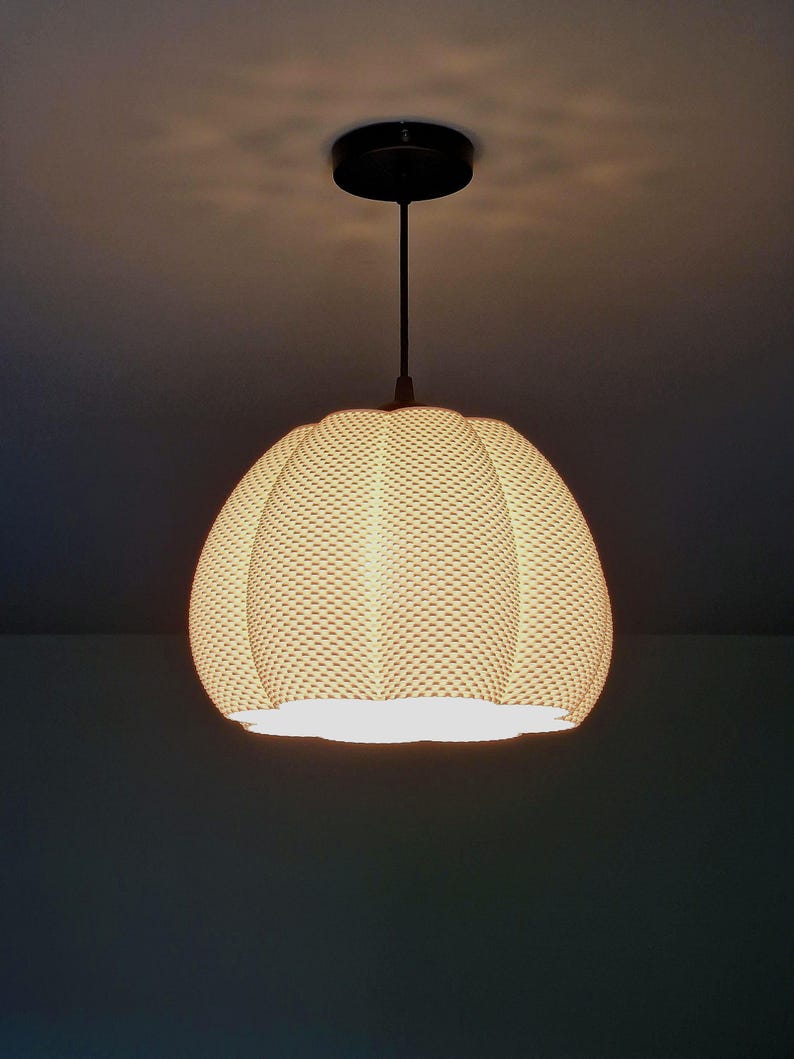 May include: A woven, beige pendant light with a rounded, textured shade. It hangs from a black ceiling mount and a slender black rod. The light is on, illuminating the room. The shade has a diameter of approximately 30 cm.