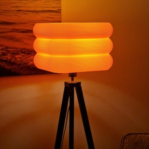 May include: A textured orange lampshade with three stacked sections sits on a black tripod lamp base. The lamp emits a warm glow. A painting of an orange sunset is visible in the background.