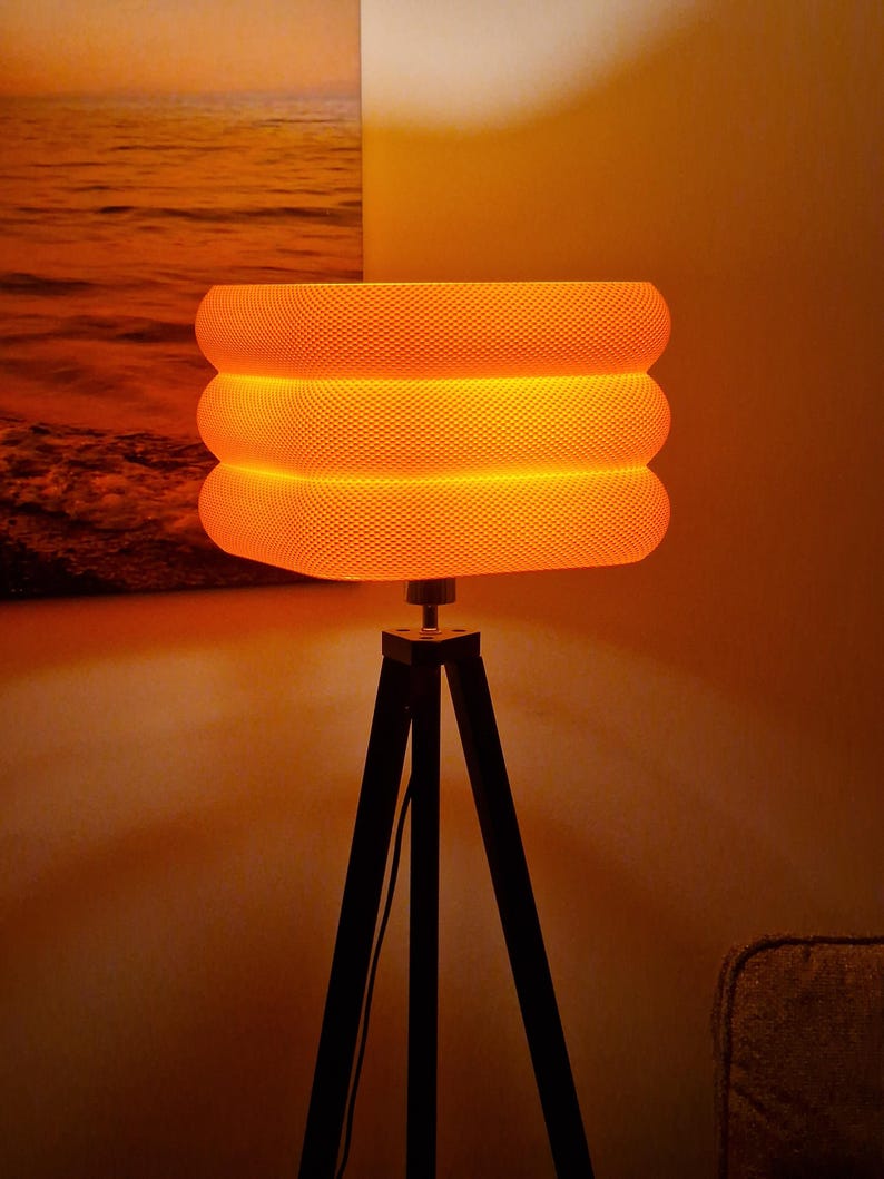 May include: A textured orange lampshade with three stacked sections sits on a black tripod lamp base. The lamp emits a warm glow. A painting of an orange sunset is visible in the background.