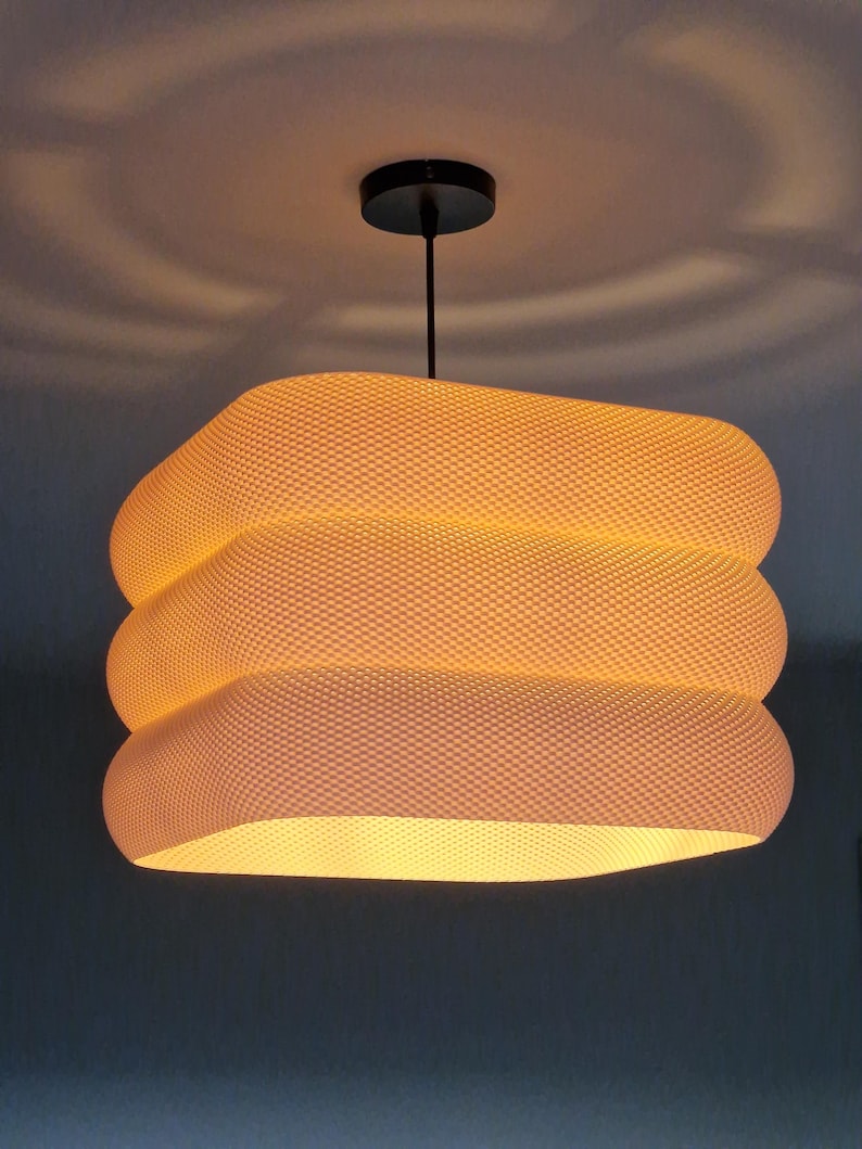 May include: A contemporary pendant light with a textured, off-white shade. The shade is square with rounded edges and is suspended from a black ceiling mount. The light emits a warm glow, perfect for a living space or dining area.