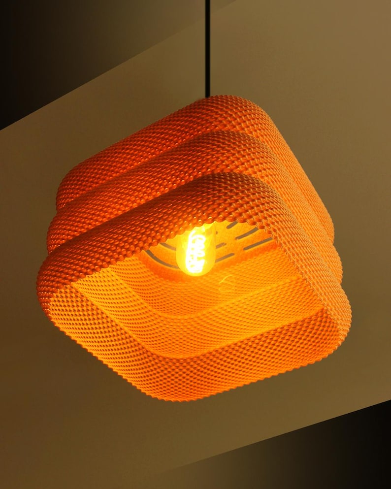 May include: Orange, textured pendant light fitting with a square shape. The light has a layered design and a visible lightbulb. The fitting is suspended from a black cord.