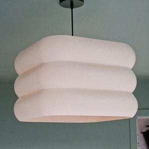 May include: A contemporary white pendant light with a textured, layered design. The light hangs from a black ceiling mount and cord. The shade has a rounded, stacked appearance, creating a unique, modern aesthetic. Suitable for indoor use, this light fixture is a stylish addition.