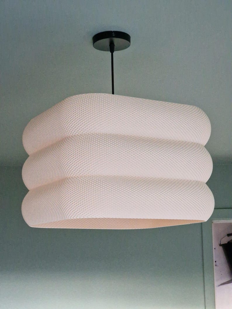May include: A contemporary white pendant light with a textured, layered design. The light hangs from a black ceiling mount and cord. The shade has a rounded, stacked appearance, creating a unique, modern aesthetic. Suitable for indoor use, this light fixture is a stylish addition.