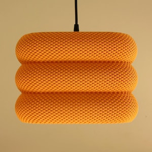 May include: An orange pendant light with a textured, woven design. The light has a cylindrical shape, comprising three stacked sections. A black cord suspends the light from the ceiling.