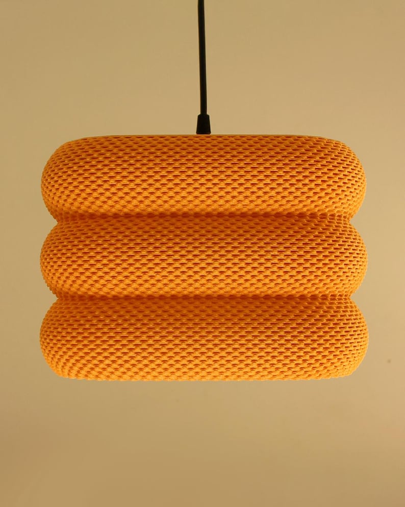 May include: An orange pendant light with a textured, woven design. The light has a cylindrical shape, comprising three stacked sections. A black cord suspends the light from the ceiling.