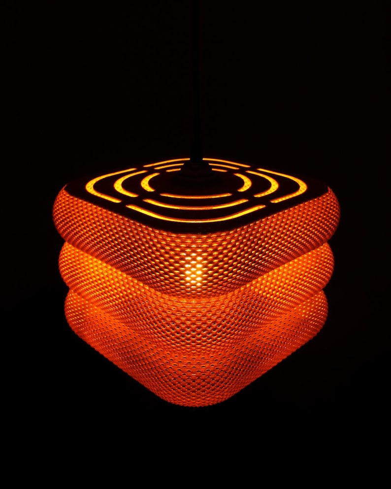 May include: An orange, textured pendant light with a unique, layered design. The light has a geometric shape with a series of concentric circles cut into the top. The light is illuminated from within, creating a warm glow against a black background.