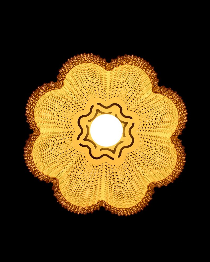 May include: A flower-shaped, yellow lampshade with a scalloped edge and a central light source. The lampshade has a woven texture and a dark brown trim, set against a black background.