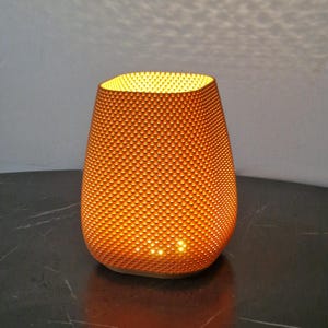 Può includere: Un portacandele decorativo arancione brillante con una superficie strutturata e puntinata. La luce brilla attraverso i piccoli fori, creando un'atmosfera calda. Il portacandele è su una superficie scura.