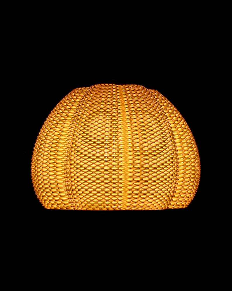 May include: A woven, dome-shaped lampshade in a warm, golden-yellow hue. The shade has a textured surface with a repeating pattern of small, rectangular openings. The background is solid black, highlighting the lampshade's shape and color.