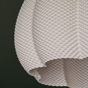 May include: Close-up of a white, textured fabric with a repeating pattern of small, raised, honeycomb-like shapes. The fabric is draped, with a visible edge and a dark background.