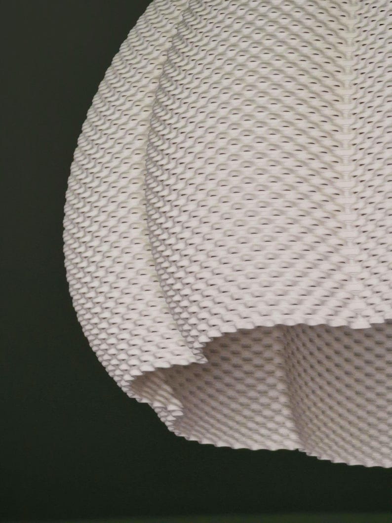 May include: Close-up of a white, textured fabric with a repeating pattern of small, raised, honeycomb-like shapes. The fabric is draped, with a visible edge and a dark background.