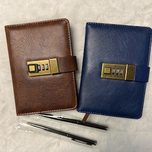 May include: Two leather-bound journals, one brown and one blue, each with a combination lock closure. Two black pens are included. The journals are designed for personal use, note-taking, or journaling.