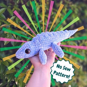 May include: A light blue crocheted whale plushie with dark brown eyes and small flippers. The whale is held in a hand, with a green, pink, yellow, and orange burst design in the background. The text "No Sew Pattern!" is in a white and green starburst.