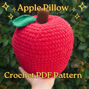 May include: A red crocheted apple pillow with a green crocheted leaf and a brown crocheted stem. The pillow is being held in a hand. The text "Apple Pillow" and "Crochet PDF Pattern" is visible in the image.