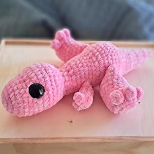 Pu&ograve; includere: Un giocattolo di peluche rosa lavorato a uncinetto a forma di lucertola con occhi neri.