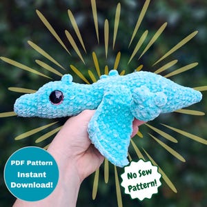 May include: A blue crocheted dragon plush toy with a large eye and a wing. The toy is being held in a hand. The text "No Sew Pattern!" is in a circle on the right side of the image. The text "PDF Pattern Instant Download!" is on the left side of the image.