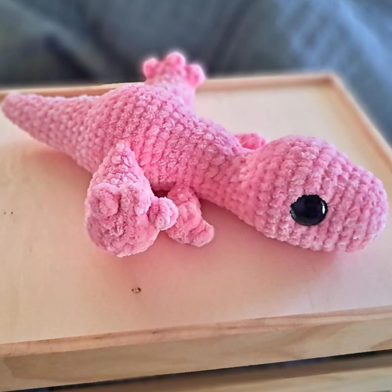 Pu&ograve; includere: Un giocattolo di peluche rosa all'uncinetto a forma di lucertola con occhi neri. Il giocattolo &egrave; sdraiato su una superficie di legno chiaro.