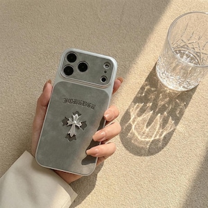 May include: A silver and gray phone case with a cross design. The case has a gray suede-like panel with the word "FOREVER" embossed above a silver cross. The phone case is held in a hand, with a glass of water in the background.