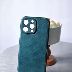 Puede incluir: Funda de teléfono de color verde azulado con un aspecto texturizado similar al cuero. La funda tiene un recorte para la lente de la cámara con círculos blancos. Es rectangular con esquinas redondeadas y un borde cosido.