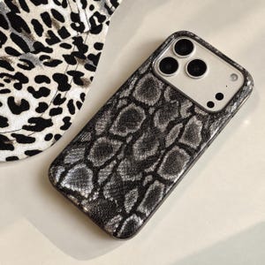 May include: A rectangular smartphone case featuring a textured black and silver snakeskin pattern. The case has a cutout for a triple-lens camera system and a flash. The surface has a metallic sheen, and the edges are finished with a dark border.