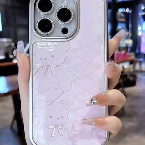 May include: A light pink phone case with a silver frame. The case features a teddy bear design and the text "Beauty and Wild". The camera lenses are surrounded by sparkling rhinestones. The case is held by a person.