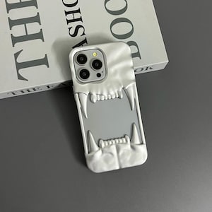 May include: A white phone case featuring a 3D design of a mouth with sharp teeth. The case is designed for a phone with a triple camera lens.