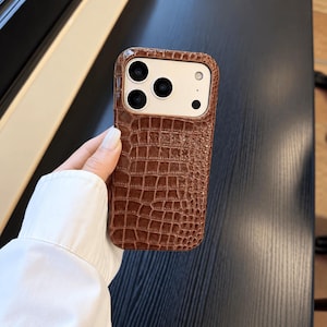 Puede incluir: Una funda para smartphone de color marrón con un patrón texturizado de piel de cocodrilo. La funda presenta un recorte rectangular en la parte superior para acomodar las lentes de la cámara y el flash, manteniendo un diseño elegante.